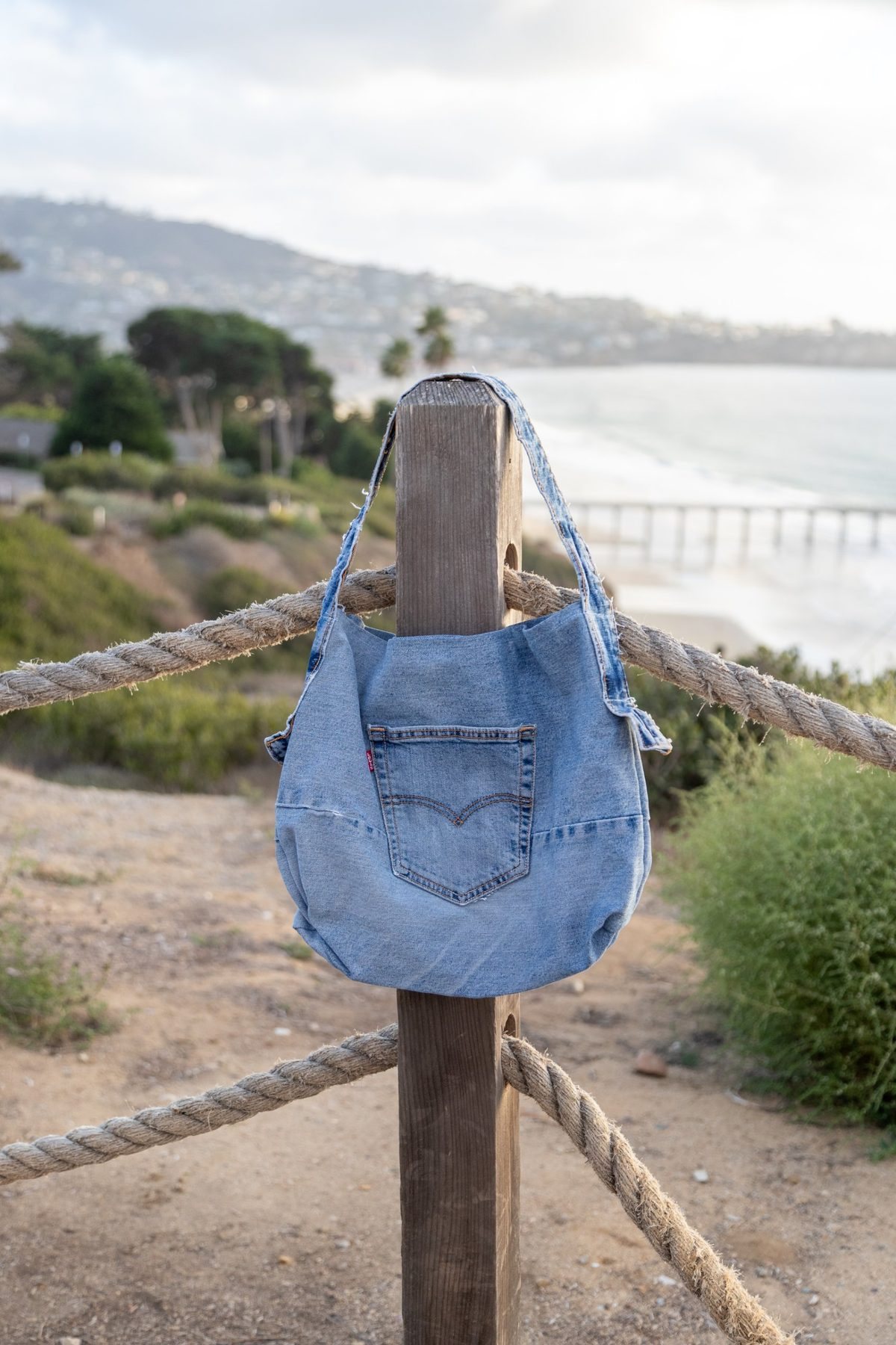 A one-of-one denim bag hanging from a rope railing above the California coast